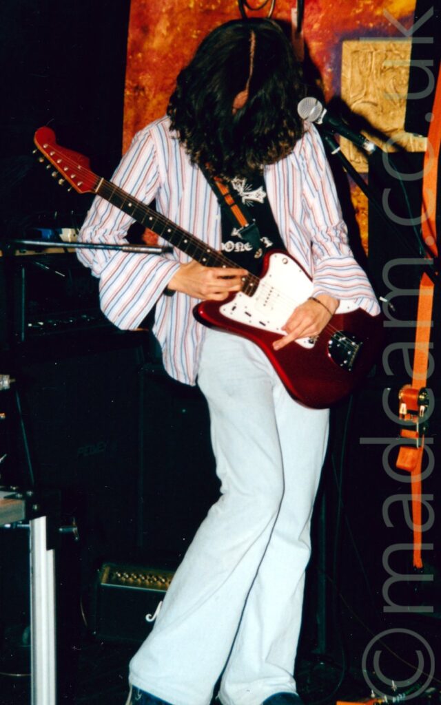 A man with long, wavy black hair, wearing an open white shirt with red and blue pin-stripes and large, open cuffs on the sleeves, over a black t-shirt with a white logo, and flared white jeans, playing a red and white guitar left-handed. He is looking down at his guitar, his face covered with his long hair. A microphone on a stand is in front of him, but he isn't using it. There are a number of black amps scattered around behind him, leaning against the orange and yellow wall, or the black wall at the back of the stage.