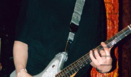 A man with short dark hair and wearing a black t-shirt and a blue pair of jeans, and playing a metallic-grey and white electric guitar. A black guitar strap, covered in grey tape, is attached to the top and bottom of the guitar, and slung over his left shoulder. He is looking down at his instrument, his mouth slightly agape. A brown, orange, and yellow pillar, looking almost organic, is behind his left arm, stretching from top to bottom of the frame. Dark walls and ceiling fill the rest of the frame, with green stagelights and yellow room lights visible here and there.