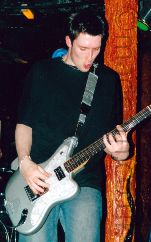 A man with short dark hair and wearing a black t-shirt and a blue pair of jeans, and playing a metallic-grey and white electric guitar. A black guitar strap, covered in grey tape, is attached to the top and bottom of the guitar, and slung over his left shoulder. He is looking down at his instrument, his mouth slightly agape. A brown, orange, and yellow pillar, looking almost organic, is behind his left arm, stretching from top to bottom of the frame. Dark walls and ceiling fill the rest of the frame, with green stagelights and yellow room lights visible here and there.