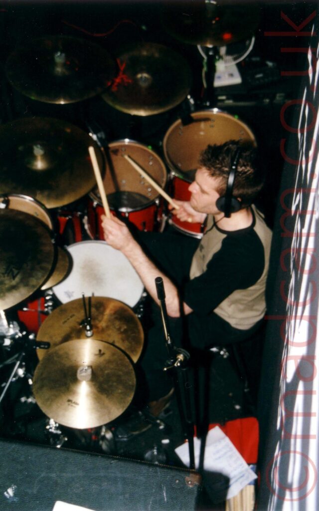 View from above of a man sitting behind a red drum kit, holding a stick in each hands as he plays. He has a pair of black headphones over his ears, red short, slightly spiky brown hair, and wearing a grey t-shirt with black sleeves.