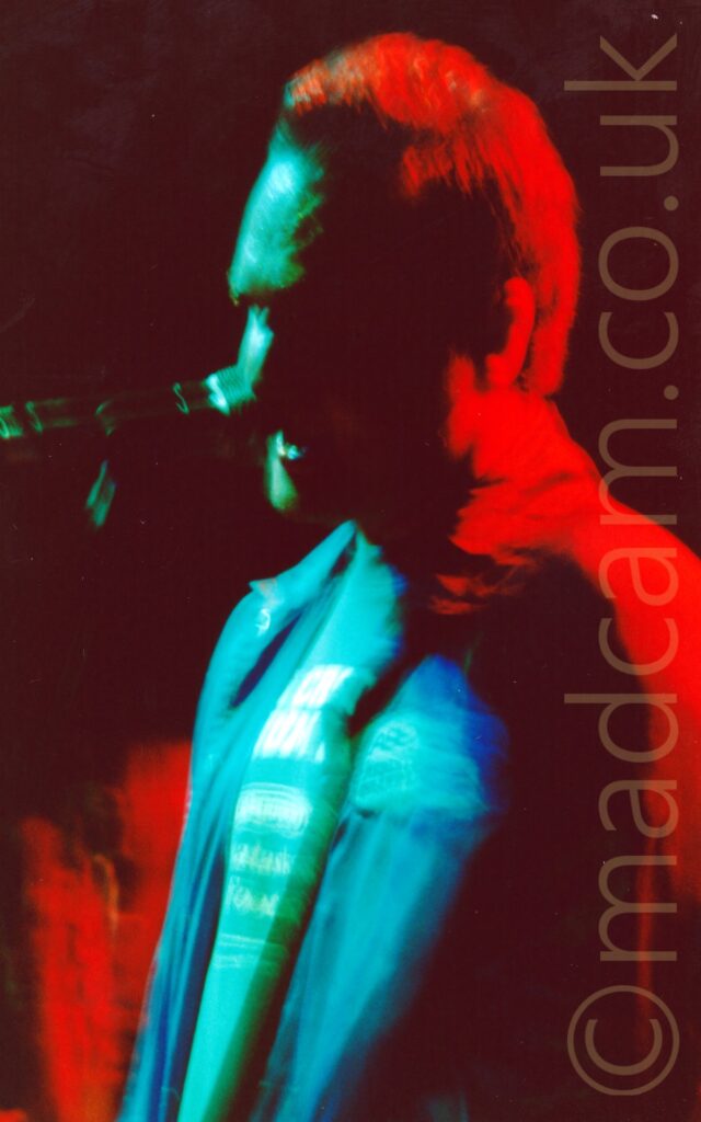 A man standing on a stage, singing into a microphone on a stand in front of him. The photo is taken with a long exposure, with stage lights flooding various parts of the frame with different vivid colours, including green on his t-shirt and face and the microphone, blue on his jacket, and red on his back and hair. Black walls fill the background.
