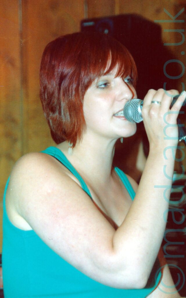 A woman with chin length red hair, and wearing a blue tank top, holding a microphone in her right hand to her mouth as she sings, looking off to the right of the frame. Varnished wood panelling fills the walls in the background.