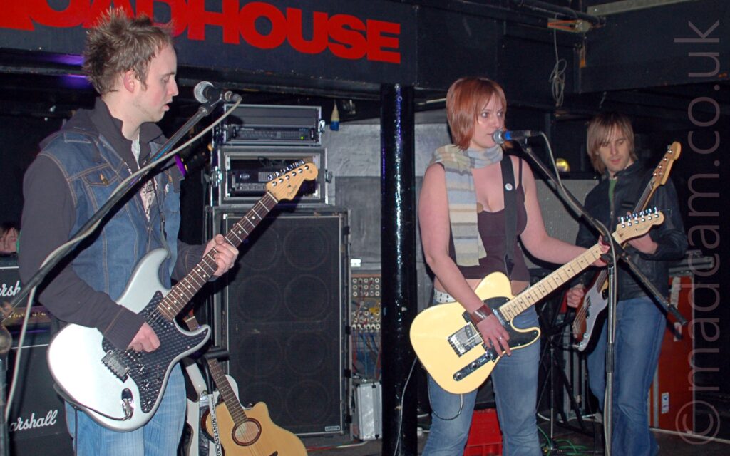 3 members of a band on a stage. On the left is a man with short, brown, spiky hair, wearing a sleeveless denim jacket over a black hoodie and blue jeans, and playing a grey and black electric guitar. In the middle is a woman with her hair cut in a brassy bob, wearing a grey woollen scarf over a strappy brown top that reveals her midriff and a pair of blue jeans, playing a yellow-ish electric guitar and singing in to a microphone on a stand in front of her. On the right is a man with shaggy brown hair, wearing a black leather jacket over a green t-shirt and blue jeans, playing a brown and white bass guitar. Racks of amplifiers line the black walls behind them, with a brown acoustic guitar casually leaning against one of the amps.
