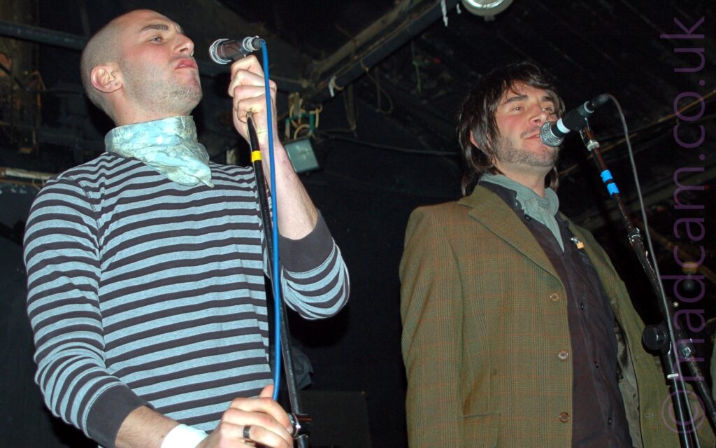 Two men standing on a stage, performing as part of a band. On the left is a man with a shaven head, wearing a grey and black striped, long sleeved t-shirt with a grey cravat round his neck, holding a microphone in his left hand and holding on to the mic stand with his right hand. On the right is a man with shaggy dark hair and a lightly-stubbly beard, wearing a green tweed jacket over a black shirt and a grey neckerchief, singing into a microphone on a stand in front of him. Black walls and ceiling fill the frame behind them.