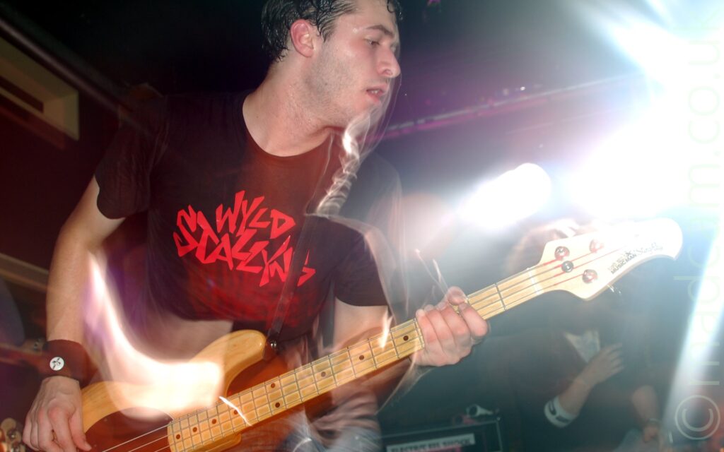 Long exposure close-up of a man with short dark hair, wearing a black t-shirt with red text "Wyld Stallions", playing a brown bass guitar. A burst of flash has frozen him in place, while his movement a snap-zoom out, and a long exposure have created secondary images, which appear to show his left shoulder being much lower than would be expected on his body. A bright white stage light on the left of the frame has created artefacts between his mouth and shoulder, as well as a bright aura around the top of his guitar. Black walls and ceiling in the background complete the image.