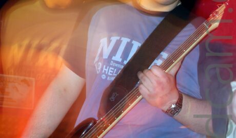 Long exposure shot of a young man wearing a blue t-shirt with barely decipherable text on the chest, and blue jeans, staring at the camera. He is laying a black bass guitar with his right hand, holding the guitars neck in his left. He has obviously moved during the exposure, with a second, orange version of him on the left of the frame in a different pose, looking down and to the right, with streaks of orange light joining the 2 images. In both images, the background is visible through his legs and body.