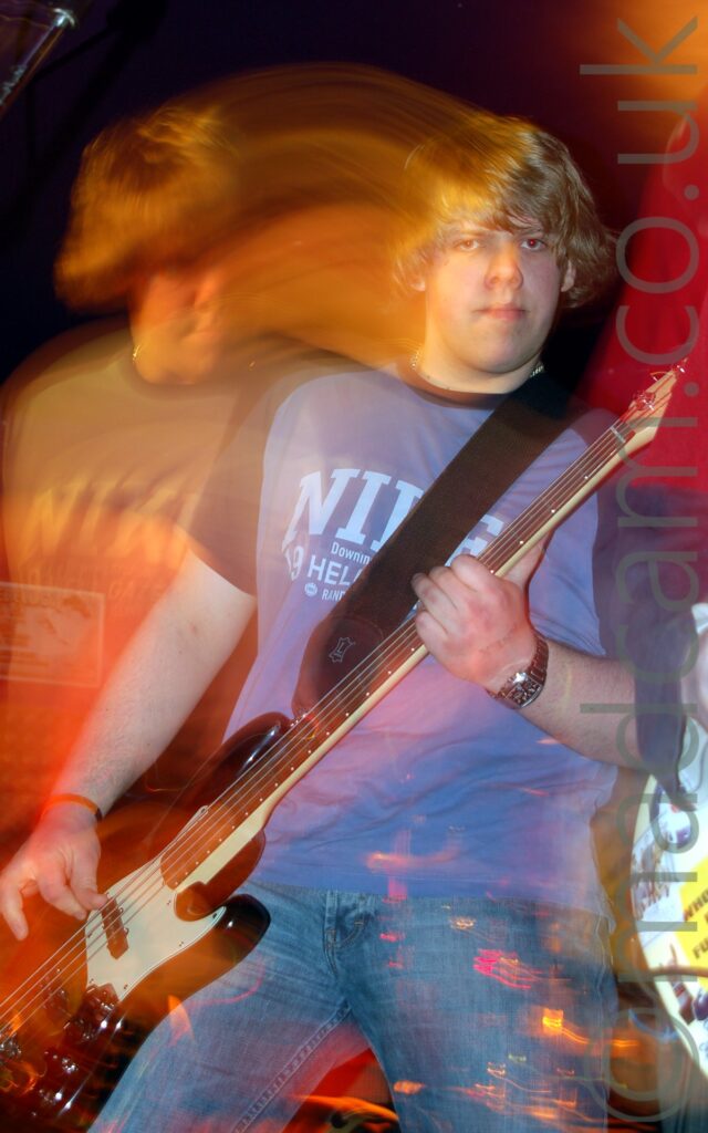 Long exposure shot of a young man wearing a blue t-shirt with barely decipherable text on the chest, and blue jeans, staring at the camera. He is laying a black bass guitar with his right hand, holding the guitars neck in his left. He has obviously moved during the exposure, with a second, orange version of him on the left of the frame in a different pose, looking down and to the right, with streaks of orange light joining the 2 images. In both images, the background is visible through his legs and body.