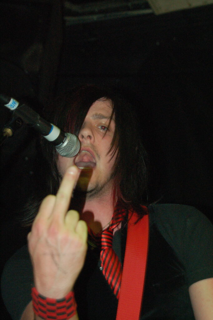 Close-up of the head and upper body of a man with long dark hair covering his right eye, wearing a grey t-shirt, a black waistcoat, and a red tie with thin black stripes, holding up an extended middle finger on his right hand to the camera, with his mouth open, about to stick his tongue out. A red guitar strap is slung over his left shoulder, and there is a microphone on a stand by his mouth on the left of the frame. Black walls and ceiling fill the rest of the frame.