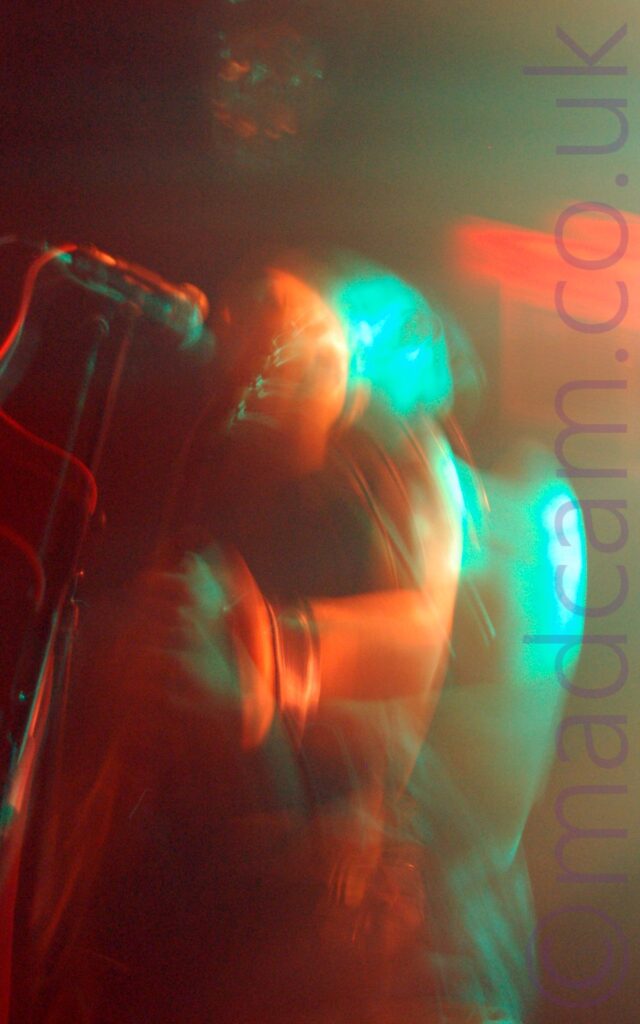 A man with a shaven head performing on stage as part of a band, playing an electric guitar and singing into a microphone on a stand in front of him. A long exposure, the stagelights, and his movements to create a red primary image of him, and a green secondary image slightly off to the right of the frame. Black walls and ceiling fill the background.