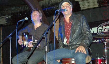 Two men sitting on stools, performing as part of a band on a stage. On the left is a man with shaggy dark hair, wearing a black shirt and black jeans, and playing a dark brown acoustic guitar, with a microphone on a stand in front of him. On the right is a mans with short blond hair, wearing a black corduroy cap, a black leather jacket over a dark red shirt with white and orange flecks, and a blue pair of jeans. He is singing into a microphone on a stand in front of his face. Behind them, black cloths cover brown brick walls, , with thick, dark brown wooden joists holding up the ceiling. A thick, tubular, black ventilation duct runs the width of the frame right at the top.