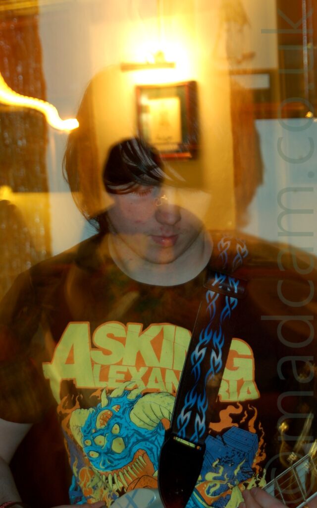 A young man with long-ish black hair, wearing a black t-shirt with a yellow, blue, and orange logo of a many-eyed, blue, demonic figure and yellow text "Asking Alexandria", and with a black guitar strap with blue flame markings slung over his left shoulder. He is looking down at the ground in front of him. A camera flash has frozen him in place, while a long exposure, a snap-zoom, and his movement have created secondary images, lit with yellow light. White walls fill the background, with a framed picture appearing in 2 different places, and wall-mounted lights that leave smears of light across the frame.