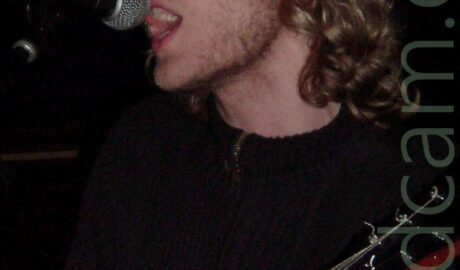 Head and shoulders shot of a man with long blond hair and stubble on his jaw, wearing a brown top, and singing into a microphone held on a stand in front of him, while playing an electric guitar. His eyes are almost completely closed. The walls in the background are almost entirely black.