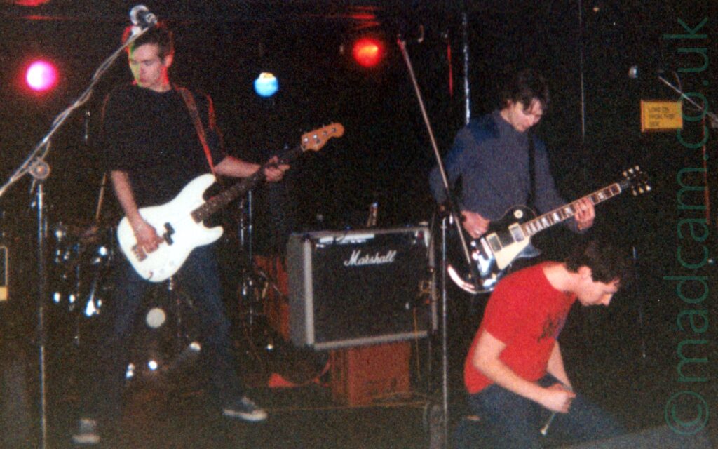 Three men on a stage, performing as part of a rock band. On the left is a man with short dark hair, wearing a black jumper and dark blue jeans, playing a white bass guitar while looking down and to his right. On the right of the frame is a man with a grey-blue top, playing a black electric guitar. In front of him, in the bottom right corner, is a man with short dark hair, wearing a red t-shirt and blue jeans, holding a microphone while kneeling on the stage. In the centre of the frame is a black amplifier with white "Marshall" text on the front in an elaborate font, resting on a red crate. Black walls and a low ceiling fill the rest of the frame, with purple, blue, and red lights shining from right at the back.