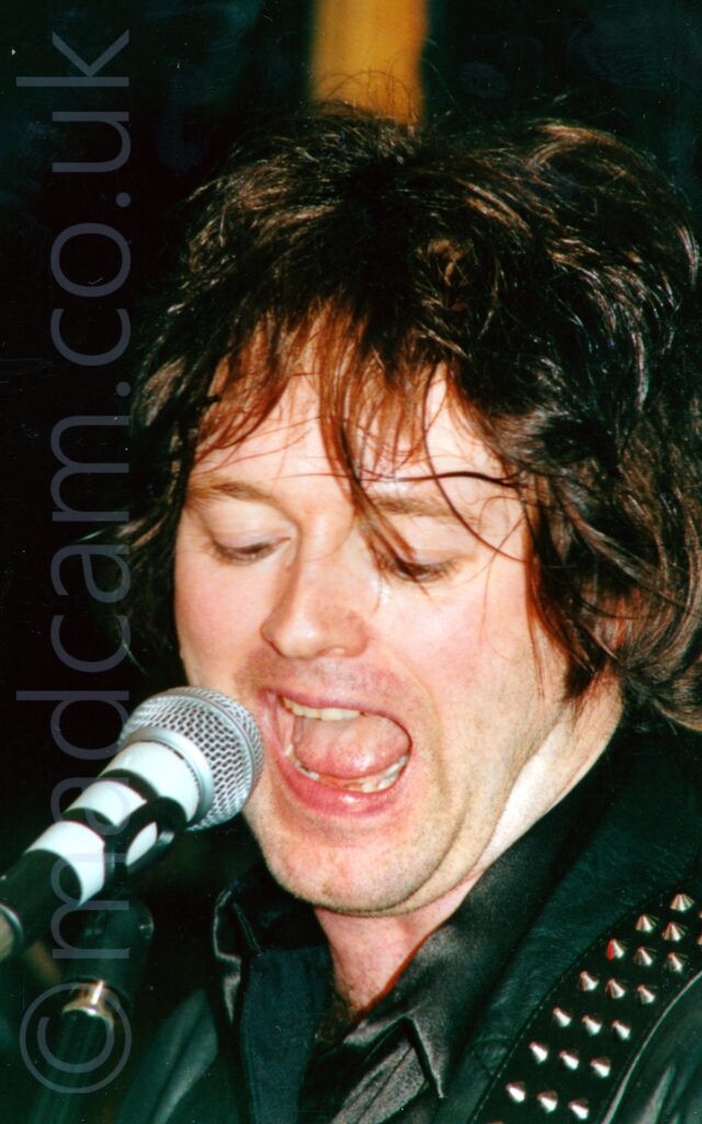 Close-up of the face of a man with dark, shaggy hair, wearing a black leather jacket over a black shirt, and with a metal-studded guitar strap slung over his left shoulder, snarling into a microphone on a stand in front of his face. Black walls and ceiling fill the background.