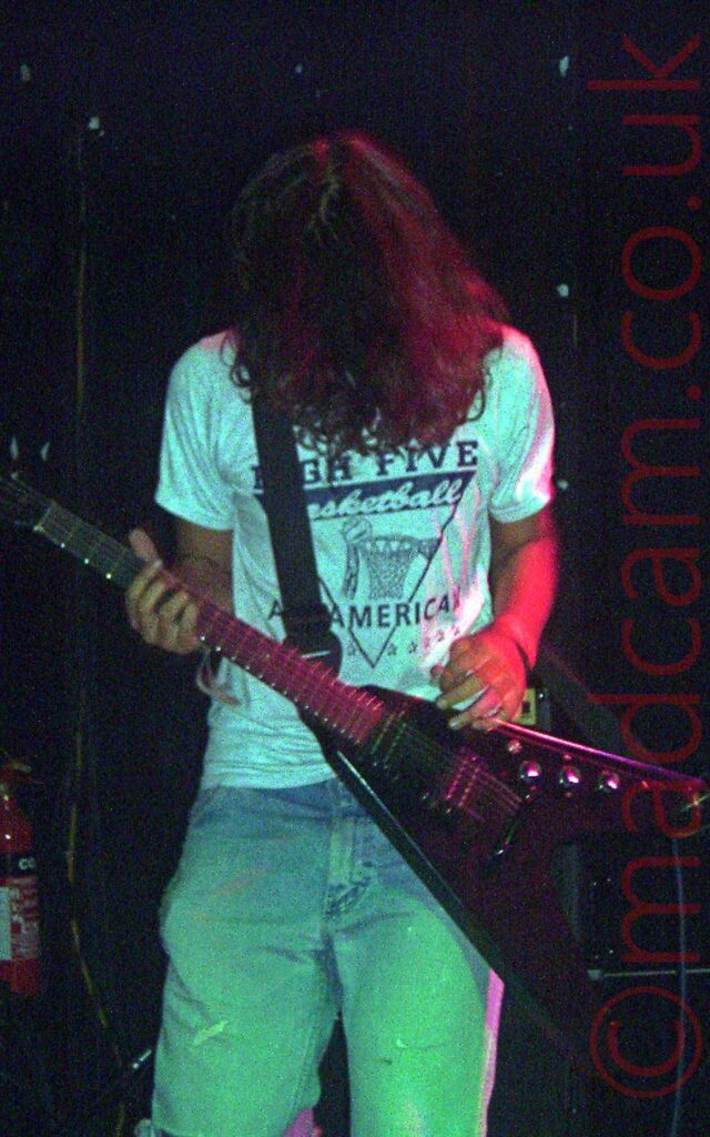 A man with long, shaggy, dark hair, wearing a white t-shirt with a black text logo and a faded blue pair of jeans, and playing a black "flying V" electric guitar, left-handed. His face is hidden in his mop of hair as he looks down at his guitar. Stage lights are giving his t-shirt a blue cast, his jeans a green cast, and his left arm is being high-lighted by a red light. Black walls and ceiling fill the background.