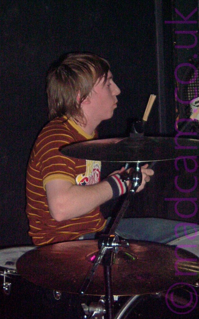 A man with shaggy chin-length brown hair, sitting behind a drum kit facing to the right, with a drum brush raised in his left hand. He is wearing a brown t-shirt with thin yellow bands around the body, and a yellow, white, and red logo on the front. A pair of large cymbals are off to his right, between him and the camera. A large black amplifier is in the background, on the right of the frame, with black walls filling the rest of the frame.