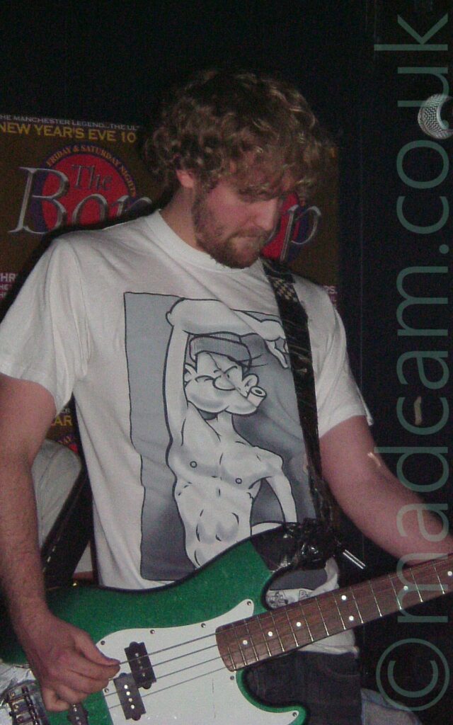 A man with wavy blond hair and a short beard, wearing a white t-shirt with a grey-scale of a muscular cartoon character, and playing a metallic green and white electric bass guitar. There is black tape holding a black guitar strap on to one of the mount points on the bass. AA brown poster on the black wall in the background has text advertising and event called "The Bop", on New Years Eve.