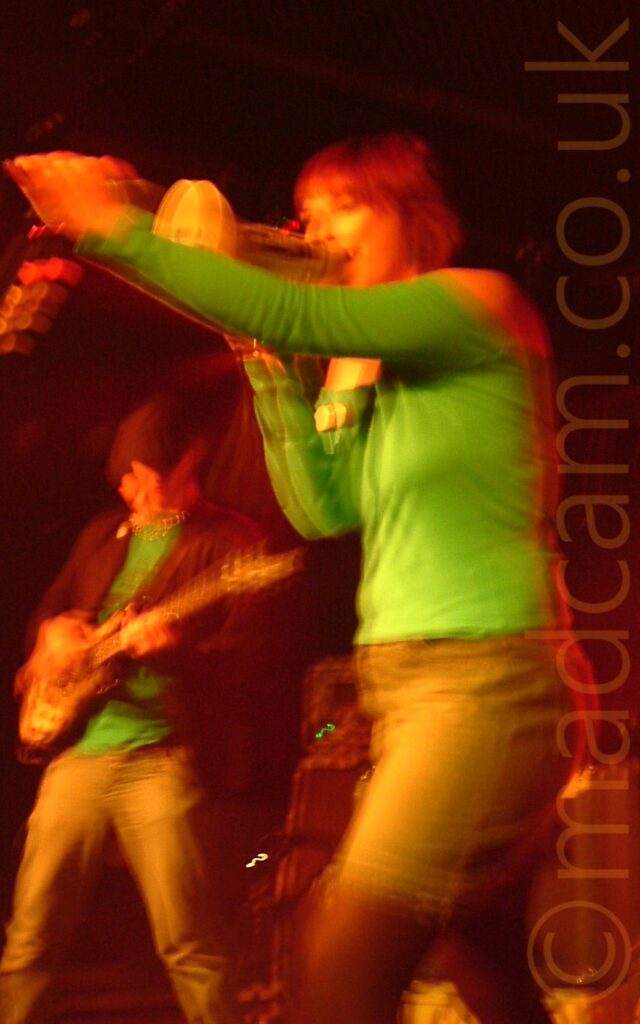 Two people on a stage, performing as part of a band. On the left, in the background, is a man wearing a black flat cap, and a suit jacket over a green t-shirt, and brown trousers, playing an electric guitar. On the right, and the main focal point of the image, is a woman with chin-length red hair, wearing a green top and a blue denim skirt, shouting into a megaphone in her right hand, held up against a microphone on a stand that she is holding with her left hand. A long exposure without a flash has created muted colours and blurred primary and secondary images, with faces and hands having an orange cast, and black walls with a red tinge filling the background.