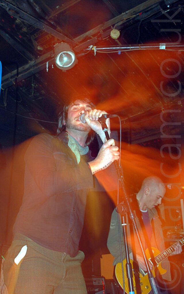 A man performing on stage as part of a band. He has chin-length dark hair and a short, stubbly beard, wearing a green neckerchief around his neck under a black shirt, and green trousers, and is holding in microphone on a stand with his left hand, his right hand wrapped around the stem of the stand, while he sings into the mic. The words "Shoes to fill" are tattooed along the side of his wrist, hand, and little finger on his right hand. A long exposure and flash, along with a snap zoom out, has created orange auras around his head, body, and arms,. In the background, in the bottom right corner, a man with a shaven head and wearing a green jacket over a blue t-shirt is playing an varnished wood electric guitar. The rest of the background is filled with black walls and ceiling, with a single blue-tinged light directly over the singers head.
