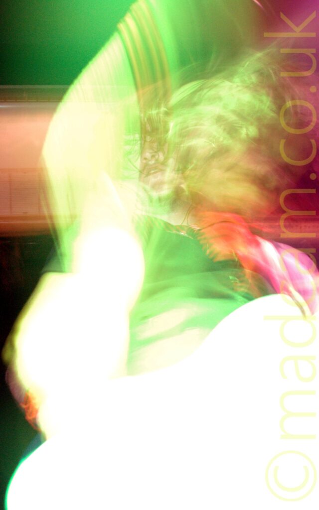 Long exposure shot of a man with long, shaggy, dark hair, wearing a black t-shirt and a loosely knotted red and black striped tie, and playing a white bass guitar. The long exposure, his movement, and the stage lights combine to leave his face perfectly focused but leaves everything else either blown out (his bass and right arm) or smeared to the point of almost being un-recognisable (his bouncing hair, left shoulder, the background). An explosion of white light has left just a vague shape where his bass and right arm should be, while green lights highlight the rest of him, and red lights highlight parts of the black wall in the background.