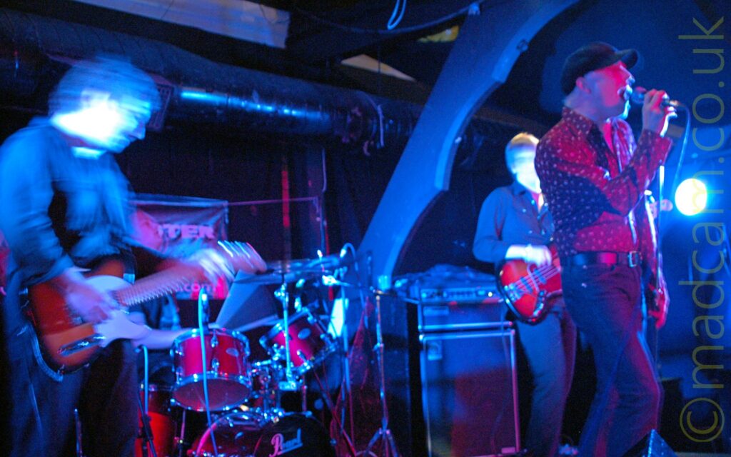 4 members of a band performing on stage. On the left is a man with short dark hair, wearing a dark top and blue jeans, playing a brown and white electric guitar. Behind him, barely visible, is a man sitting behind a red drum kit. On the right is a man with blond hair covered by a black cap, a dark shirt with light dots, and a blue pair of jeans., singing in to a microphone he holds in his right hand. Behind him is a man with short hair, wearing a dark shirt and trousers, playing a red bass guitar. Large wooden beams hold up the sloping roof, with black ventilation ducting suspended underneath. The whole image has a blue tint with purple highlights, with a bright blue light in the background on the right of the frame. A long exposure with no flash and the various members of the band moving mean that everything is slightly blurred, with some secondary images, particularly on the guitarist.