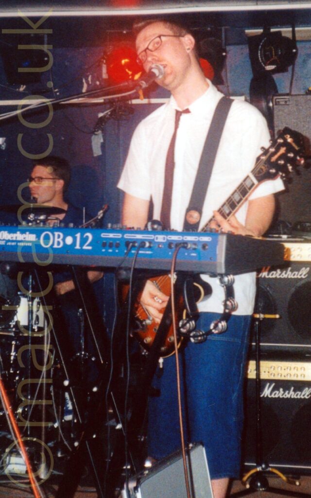 A man with short dark hair, with a pair of glasses on his face, wearing a white shirt with a brown tie and a pair of wide denim long shorts. He has a brown electric guitar slung over his shoulder, which he is playing while singing into a microphone on a stand off to his right. He is standing behind a blue keyboard with white "Oberheim OB2" text on the back edge. A pair of Marshall amps are stacked behind him on the right of the frame, with a man behind a drumkit on the left, with purple walls and black ceilings right at the back.
