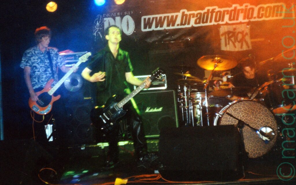 3 people on stage, performing as part of a rock band. On the left is a man with shaggy, ear-length brown hair, wearing a blue and grey shirt, and playing an orange bass guitar. In the middle is a man with short dark hair, wearing a black shirt and dark trousers, playing a black electric guitar. On the right is a man with short dark hair, sitting behind (and playing) a black drum kit at the back of the stage. The wall at the back is black, with large black text "www,bradfordrio.com" outlined in white, with a smaller, white sign below with the black word "Trick". There are different coloured lights aimed at each person on stage - the bassist is highlighted in blue, an acid green glow surrounds the guitarist while a bloom of orange light surrounds the drummer.