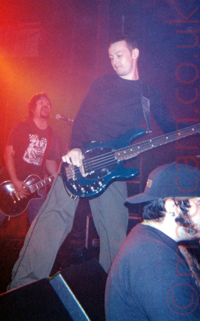 A man standing on a stage, wearing a black jumper and grey trousers, playing a blue bass guitar, and staring down and to his right. In the background, on the left, is a man with shaggy dark hair and beard, wearing a black t-shirt with a large white logo, and playing a black electric guitar while singing into a microphone on a stand in front of him. Red and orange light floods the stage on the left, with black walls on the right of the background. In the foreground, a man wearing a backwards black baseball cap and a black t-shirt, facing away from the stage, leaning back against wedge speakers on the front of the stage.