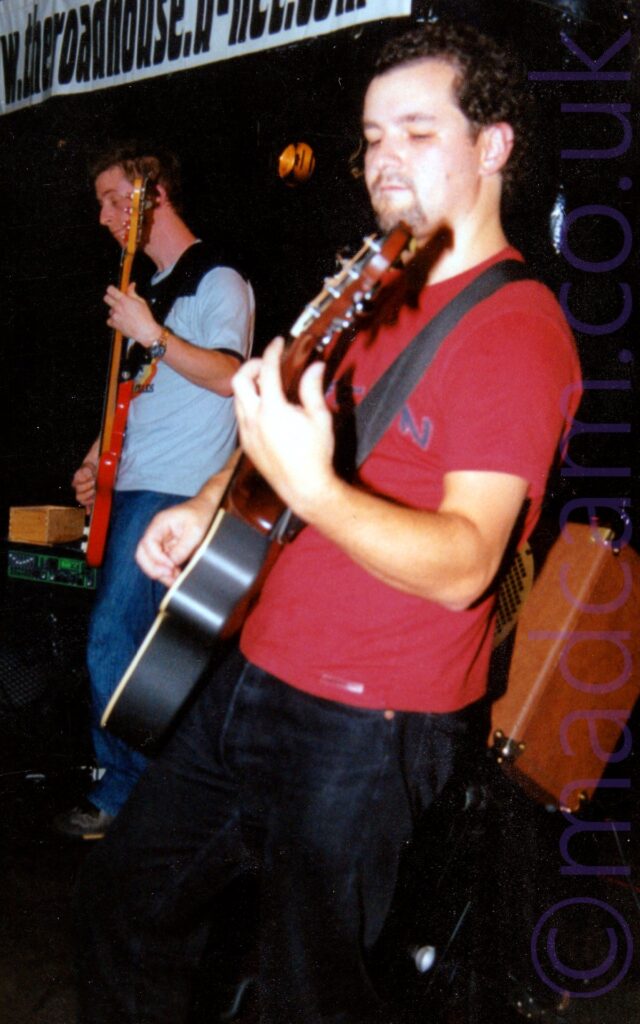 A view from the side of a man with short, curly dark hair, wearing a red t-shirt and a black pair of jeans, and playing a black acoustic guitar, looking down at his left hand as he stands facing off to the left of the frame. An orange speaker is mounted at an angle behind him, while a taller man with short dark hair and wearing a light and dark blue t-shirt is playing a red bass guitar off to the left of the frame. Black walls and ceiling fill the rest of the frame.