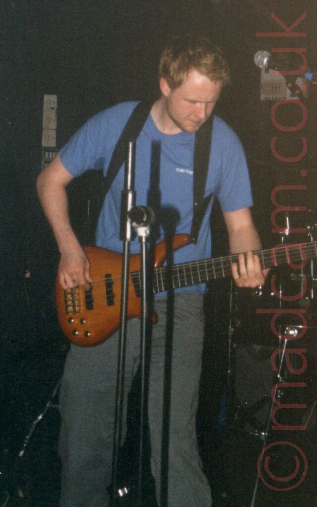 A man with short brown hair, wearing a blue t-shirt and a grey pair of trousers, playing a brown bass guitar that is supported on a harness around his neck rather than the usual strap. A microphone stand with no microphone is in front of him, while the background is black and grey amplifiers against a black wall.