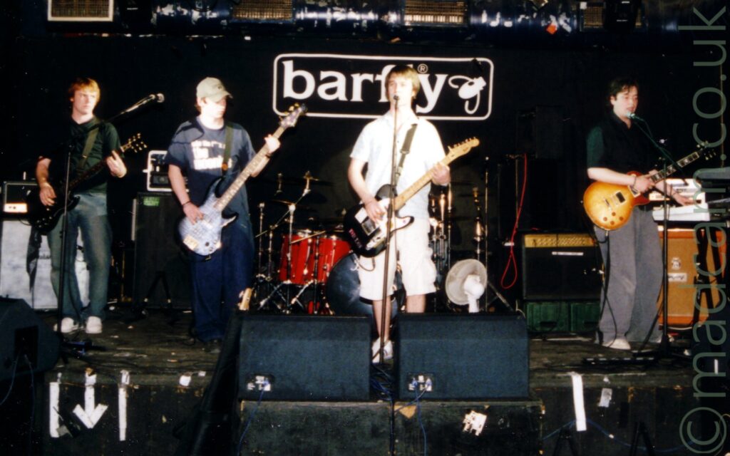 4 members of a band standing on stage playing their instruments, and 2 of them singing. On the left is a man with ginger hair and wearing a black shirt and blue jeans, playing a black guitar and looking to his left. Towards the centre-left of the frame is a man with brown hair under a green baseball cap, wearing a black t-shirt with the words "Who the f**k are the moonies" and dark blue jeans, playing a dark blue bass guitar, and looking to his left. In the centre of the image, is a man with brown hair, wearing a white shirt and white knee-length shorts, playing a black electric guitar, and standing behind a microphone stand - although his mouth is hidden by the microphone, he is singing. On the right is a man with dark hair and wearing a black shirt and flared grey jeans, holding a golden orange electric guitar in his right hand, playing keyboards with his left hand, and singing into the microphone in front of him. There is a red and chrome drum kit behind the singer in the middle, and speakers facing the stage at the front. The back of the stage is covered in a black curtain with the word "barfly" and the outline of a large fly printed on it, all in white, and outlined with a large rounded rectangle.