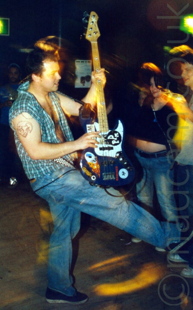 A man facing to the right while playing a blue bass guitar covered in colourful stickers, and getting up close and personal with people in the audience, his right leg extended out to the front.. He is wearing a blue and cream checked shirt, open to the navel, with the sleeves cut off, and blue jeans. The 2 people closest to him, a woman wearing a black top and blue jeans that expose her midriff, and a man wearing an open grey shirt, seem to be having different experiences - he is loving it, she doesn't seem impressed with the bassists antics. More people can be seen in the background. A flash has frozen them all in place, while a long exposure and slight zoom creates streaks of golden light and secondary images as they all move. Black walls fill the rest of the frame.