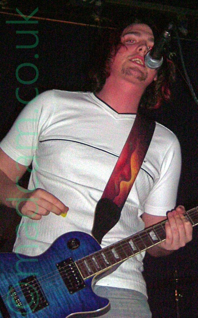 A man performing on a stage as part of a rock band. He has floppy long hair, and is wearing a white t-shirt over blue jeans, and has a blue electric guitar slung around his neck on a long black strap. He is singing in to a microphone on a stand in front of him as he plays his guitar. Black walls and ceiling fill the rest of the frame.