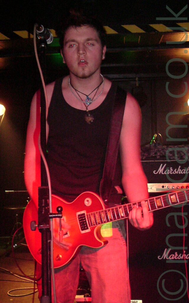 A man with spiky, short, dark hair, and wearing a black vest and dark jeans, playing an orange electric guitar, and looking somewhat tired as he stares at a microphone on a stand in front of him. A black and yellow stripe marks a low ceiling on an alcove behind him, with yellow lights in the alcove illuminating a couple of patches on walls and floor, the rest hidden in darkness.