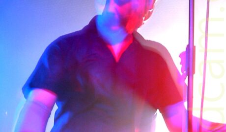 A man with a short dark hair, wearing a black shirt and trousers, and playing a bass guitar. A microphone on a stand is in front of his face. A long exposure and his movement has him looking forward, and down at his instrument, at the same time, while a slight snap zoom and stage lights creates blue, red, and purple auras around everything, with an explosion of white light backlighting the whole thing.