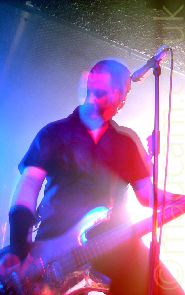 A man with a short dark hair, wearing a black shirt and trousers, and playing a bass guitar. A microphone on a stand is in front of his face. A long exposure and his movement has him looking forward, and down at his instrument, at the same time, while a slight snap zoom and stage lights creates blue, red, and purple auras around everything, with an explosion of white light backlighting the whole thing.