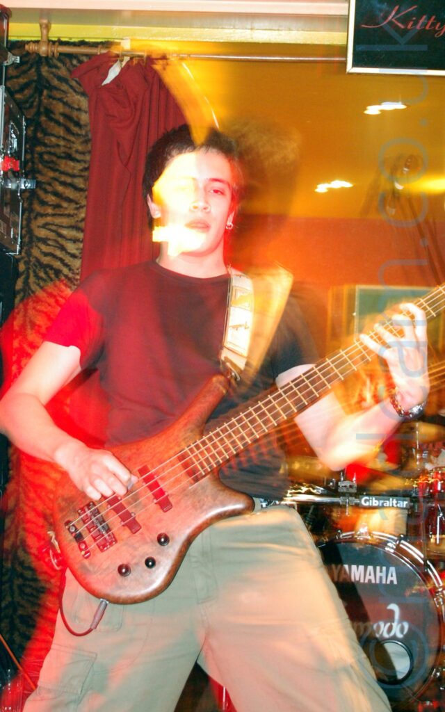 Long exposure of a man with short dark hair and wearing a black t-shirt and beige trousers, playing a brown bass guitar and looking directly at the camera. A flash has frozen him in place, while a long exposure gives the impression of movement, with red and yellow secondary images of him painted to either side. A drum kit is visible behind him in the bottom right of the frame. Red and tiger print drapes cover the walls, while the ceiling is yellow.