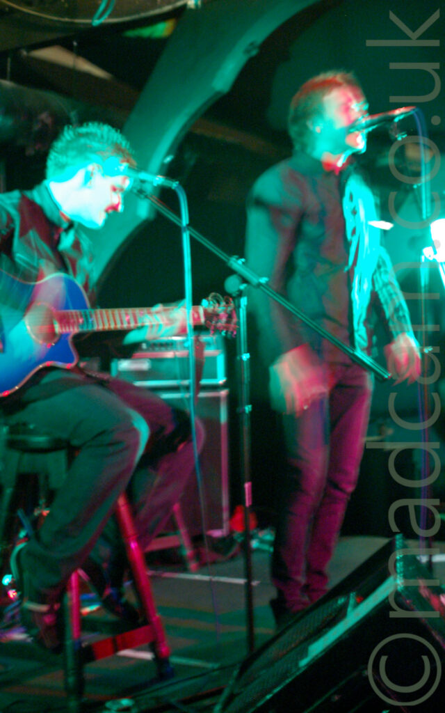 2 members of a band performing on stage. On the left, a man with short spiky hair, wearing a denim jacket and dark coloured trousers, sitting on a 4-legged wooden stool while playing a blue acoustic guitar. On the right is a man with short dark hair, wearing a light jacket and blue jeans, standing in front of a microphone on a stand as he sings. Green stage lights flood the entire frame, with wooden frames supporting the roof in the background.