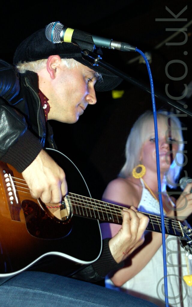 Close-up of a man with short blond hair, wearing a black corduroy cap, a leather jacket, and a blue pair of jeans, sitting on a stool and playing a black and brown acoustic guitar, facing to the right. A microphone on a stand with a blue cable dangling down is in front of his face. In the background, a woman with bleach-blonde hair cut in a long bob and wearing a strappy white dress is slightly out of focus. Black walls and ceiling fill the rest of the frame.