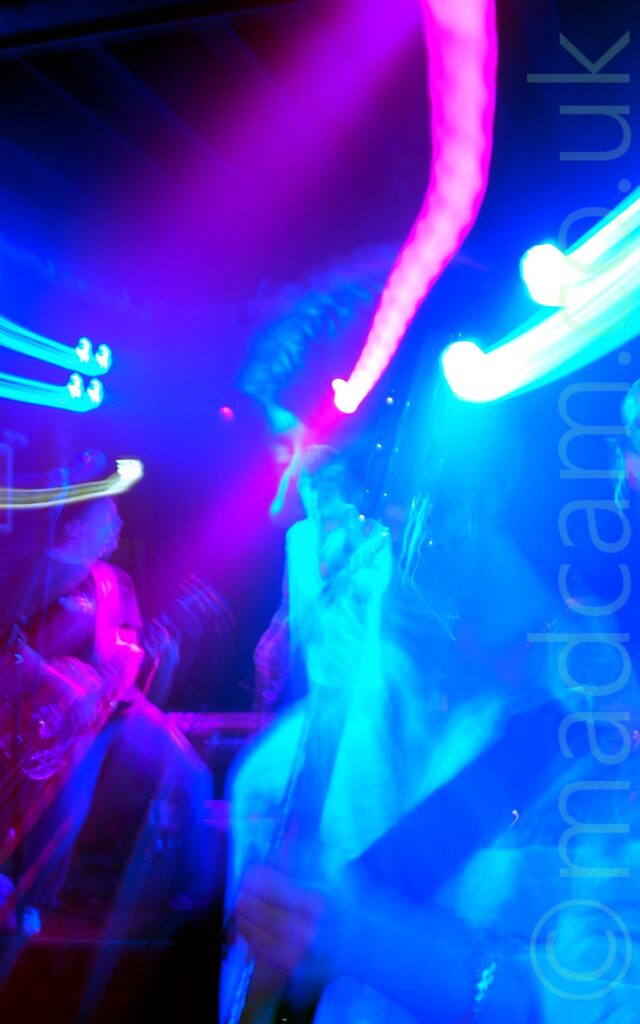 Close-up shot of a man with mid-length dark hair and wearing a light-coloured shirt, playing a bass guitar. A long exposure and a snap zoom means he appears twice in the photos, bathed in blue light, the second time appearing far away. On the left of the frame, another member of the band is playing an electric guitar, illuminated by a purple light. Streaks of blue and purple light flood the top half of the image.