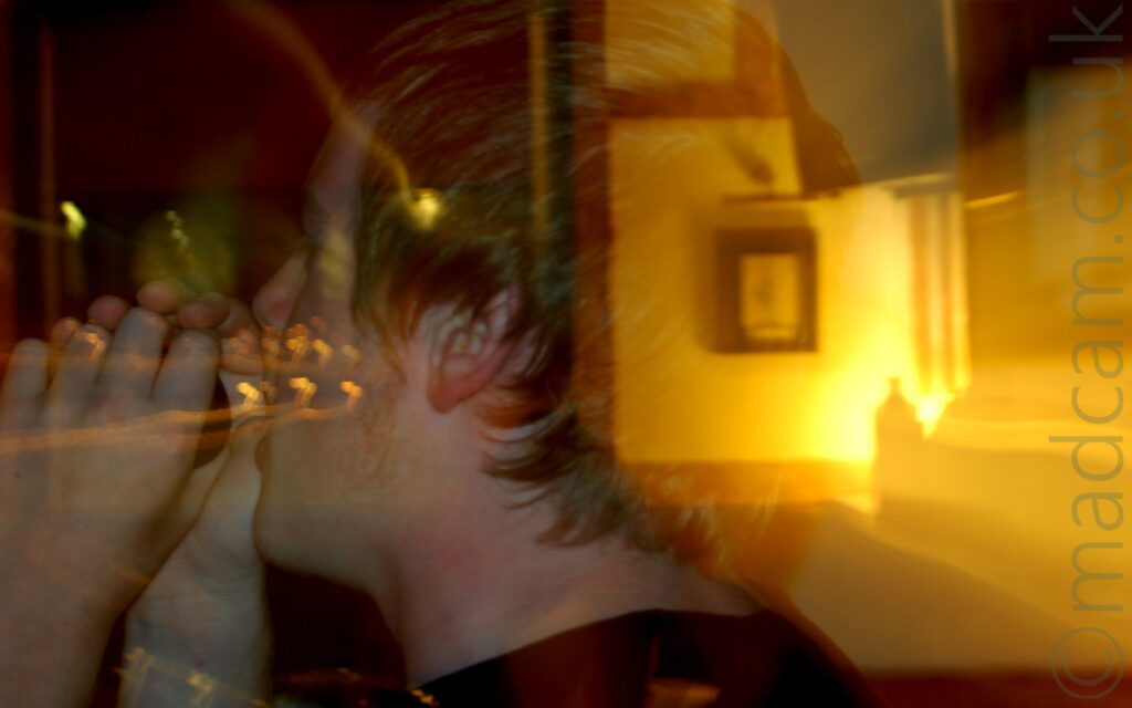Long exposure close-up of the side of a young man's head facing to the left. He has short blond hair, and is wearing a black t-shirt in his hands which is raised to his mouth as he sings into it. A snap zoom has been used, starting zoomed right in, then zooming out, meaning that the background, including a picture in a frame on the wall, appears to be visible through the back of his head, lit by yellow light. Yellow light trails are visible in the dark background on the left of the frame.