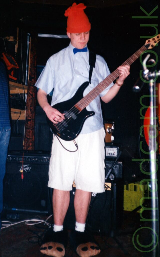 A man wearing a red woollen hat on his head, wearing a short-sleeved blue shirt with a dark blue bow tie, and long white short trousers. He has novelty slippers resembling large animal feet on his feet, and is p[laying a black electric bass guitar. Black amplifiers and speakers are in the background, along with yellow crates and wooden shelves, with black walls filling the rest of the frame.