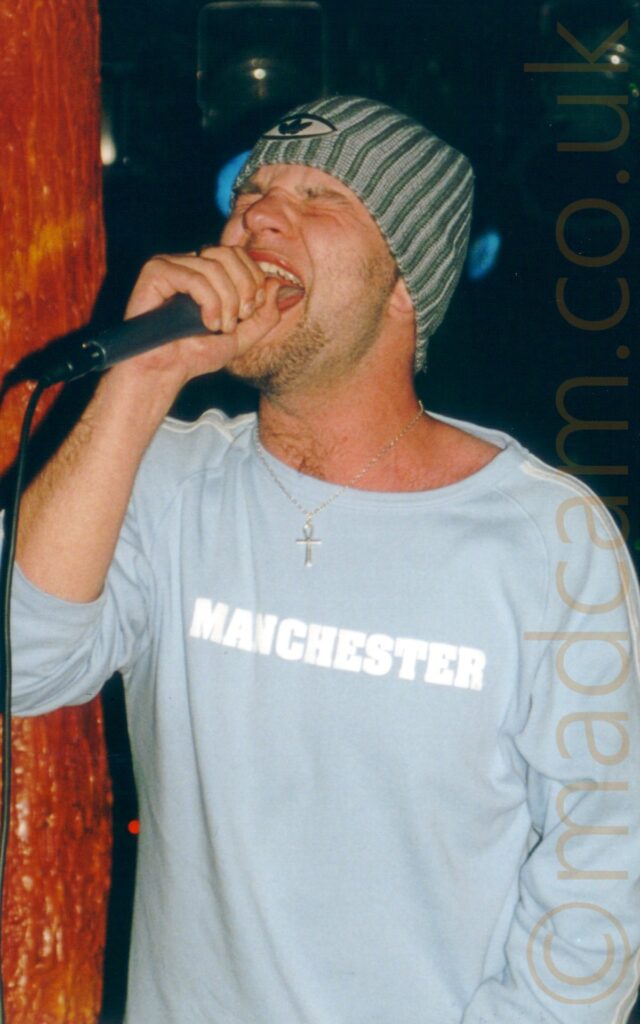 A man wearing a grey woollen hat, and a long-sleeved pale blue s-shirt with white text "Manchester" across the chest, as he screams into a microphone held in his right hand, with his eyes closed. A viscous, moist, and organic-looking amber pole fills the frame on the left, with black walls filling the rest of the background.