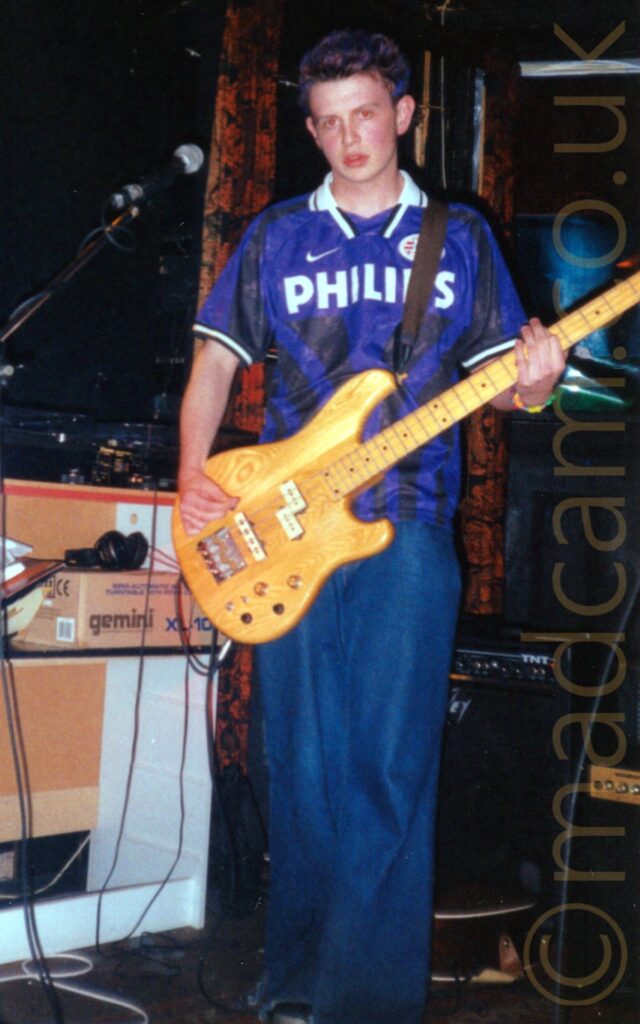 A man on a stage, with blue highlights in his short brown hair, wearing a blue and black sport shirt with the large white word "Phillips" on the front, and baggy blue jeans. He is playing a varnished wooden bass guitar, and staring towards the camera. Cardboard boxes line the walls in the background on the left, With amplifiers on the right, and brown curtains covering the wall in the centre.