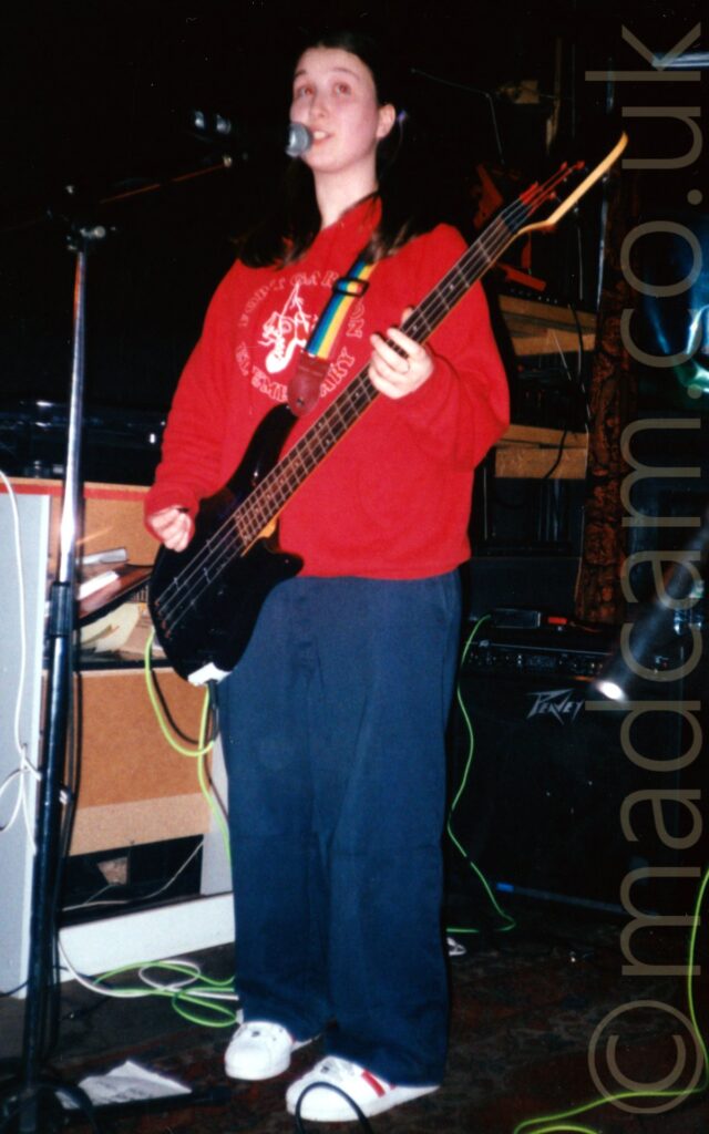A woman with long dark hair cascading over her shoulders, wearing a red hoodie with a circular white logo ion the front, and a blue pair of baggy jeans, playing a black bass guitar, and singing into a microphone on a stand in front of her, while looking wistfully off into the distance. A rainbow-striped guitar strap is attached to her bass, slung over her left shoulder. Wooden shelving and black guitar amps fill the background, with black walls filling the rest.