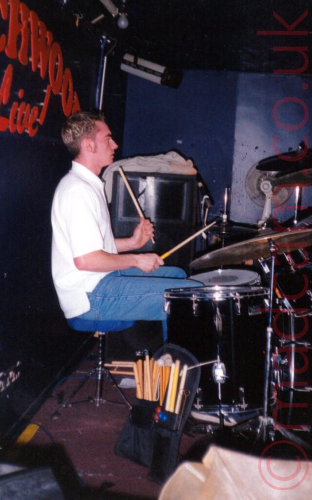 A man with short dark hair, wearing a white shirt and a blue pair of jeans, sitting on a blue drum stool behind a black drum kit, his back almost touching the wall behind him. He is holding a stick in each hand, raised slightly in the air. A bag holding more sticks is on the floor in the foreground, leaning against one of his drums. The dark blue wall on the left has red text , partially reading "chwood Live" above his head. Dark blue walls and ceiling fill the background.