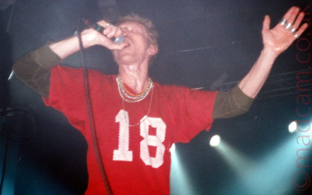 A man with spiky brown hair, wearing a red t-shirt with the white number "18" emblazoned across the front over a brown sweater, with several gold and other metal chains around his neck, holding a microphone in his right hand as he sings, with his head tilted back. Metal rings are around each of the fingers in his outstretched left hand. Black walls and ceiling fill the background, with 4 white lights shining in the bottom right corner.