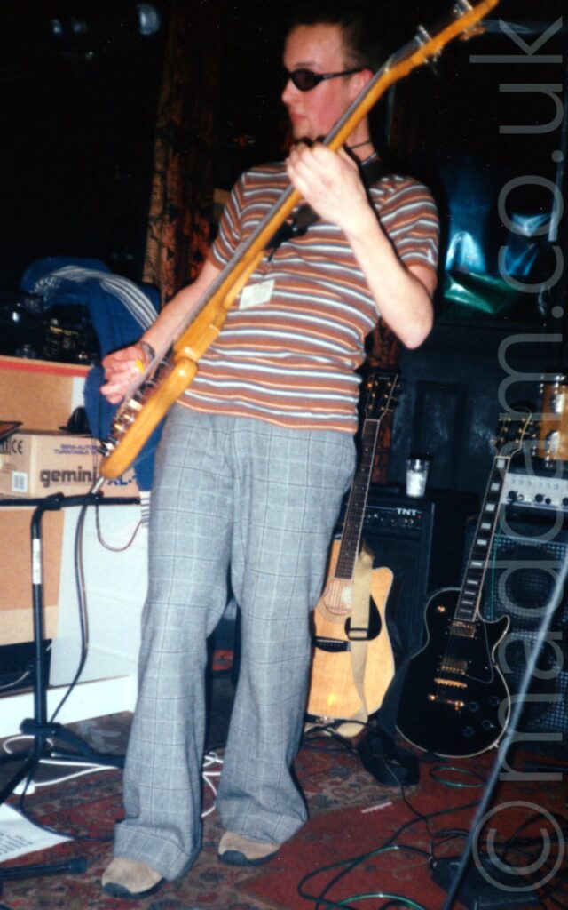 A man with short dark hair and wearing a dark pair of glasses, wearing a brown, grey, and white stripey shirt and grey trousers, and playing a polished wooden bass guitar on a stage. Assorted guitar stands, cardboard boxes, guitars, and amplifiers line the side and back of the stage, with black walls filling the rest of the frame.