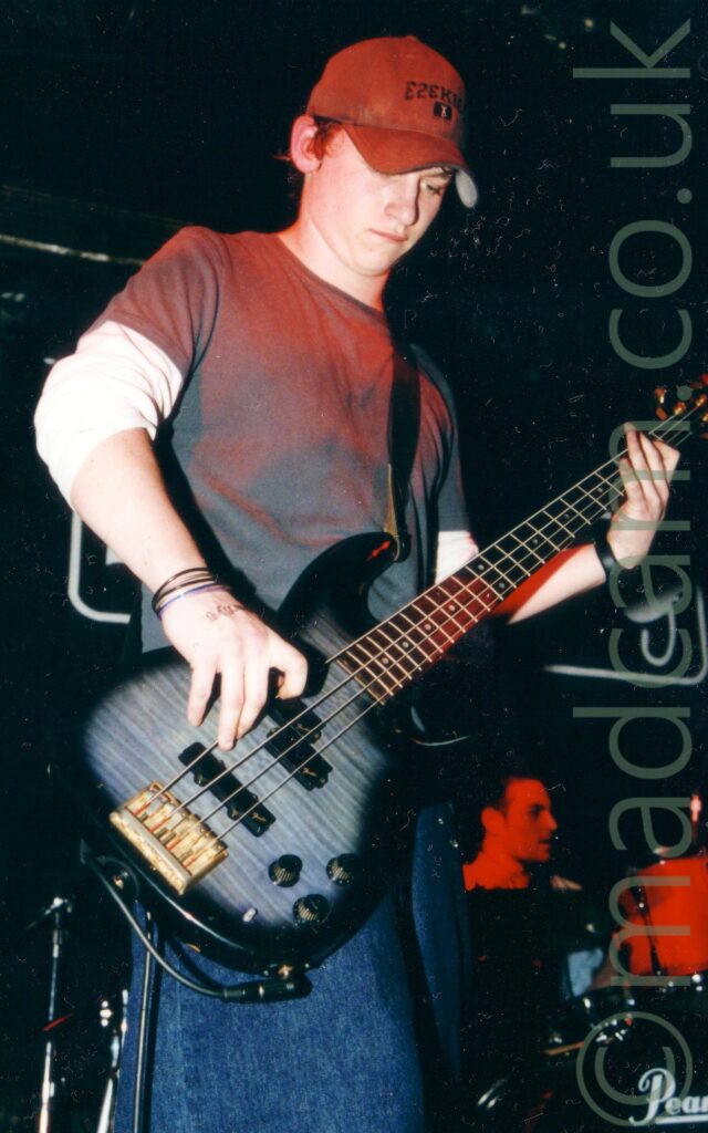 A man wearing a grey baseball cap with black text "Ezekiel" on the front, and a grey t-shirt over a white long-sleeved t-shirt with the sleeves rolled up to just below his elbows, and a blue pair of jeans. He is playing a black and grey bass guitar, and looking down towards the floor. A man can be seen sitting behind a drum kit in the bottom right corner, bathed in red light from the stage lights. Black walls and ceiling fill the rest of the background.