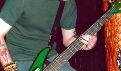 A man with short dark hair and beard, wearing a green t-shirt and a grey pair of cargo pants, playing a bright green 5-stringed bass guitar. His right wrist is covered in an array of beaded bracelet, while his right forearm has a tattoo of a red love heart with a large knife through it. His left forearm has a black tattoo which is partially hidden behind the neck of the bass. He is slightly hunched over, looking down at his left hand. In the background, an orange pole fills the right of the frame, glistening wetly, with a visceral, organic appearance, with black walls and ceiling filling the rest of the frame.
